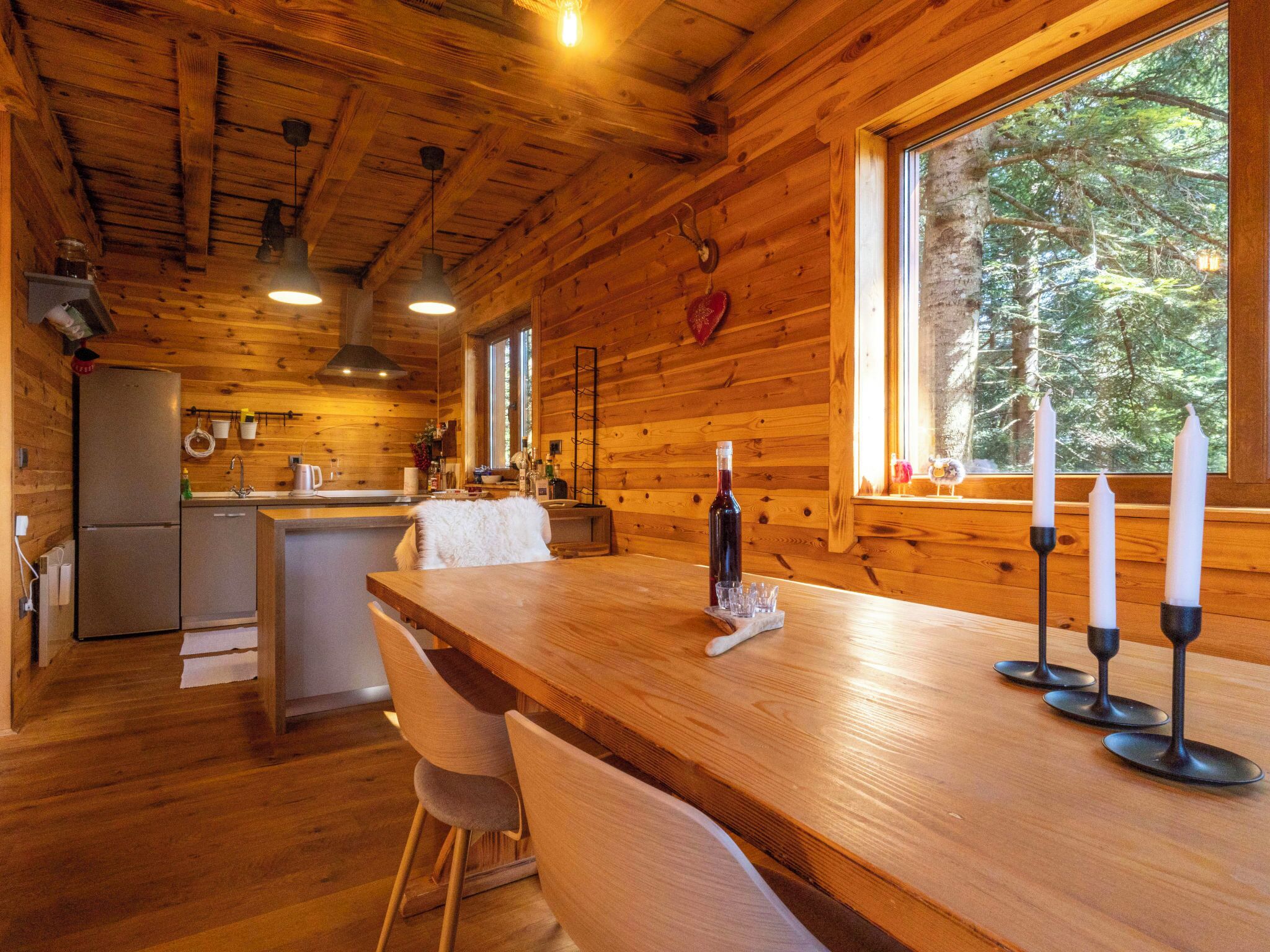 A wooden cabin interior with a kitchen, dining area, and large windows.