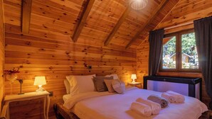Quarto de cabana de madeira com uma cama, duas luminárias de cabeceira, uma janela com cortinas e uma pequena mesa com abajur.