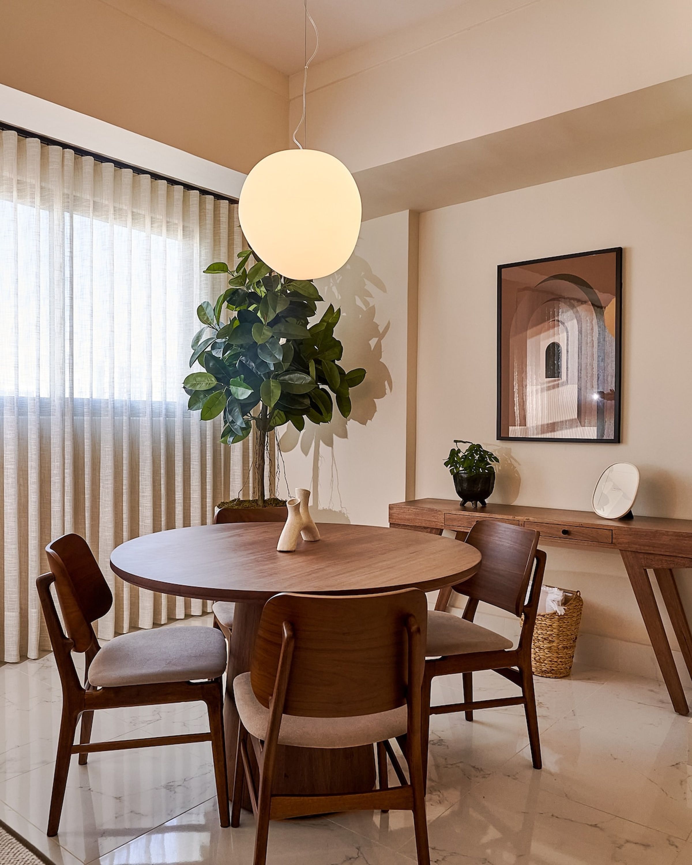 Een eetgedeelte met een ronde houten tafel, vier stoelen, een hanglamp, een potplant, een lijst met een afbeelding aan de muur, en een houten bureau met een mand.