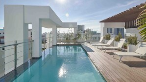 En pool på taket med fri utsikt över stadens silhuett, omgiven av vita väggar och solstolar.