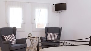 Une chambre à coucher avec deux fauteuils, un lit, un téléviseur fixé au mur et une petite table sur laquelle se trouvent des livres et une horloge.