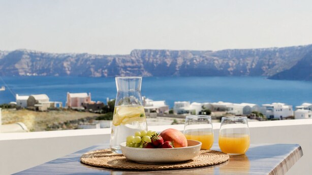 Une table avec une carafe d’eau, un saladier de fruits et des verres de jus d’orange, donnant sur un paysage côtier avec des bâtiments blancs et des falaises.