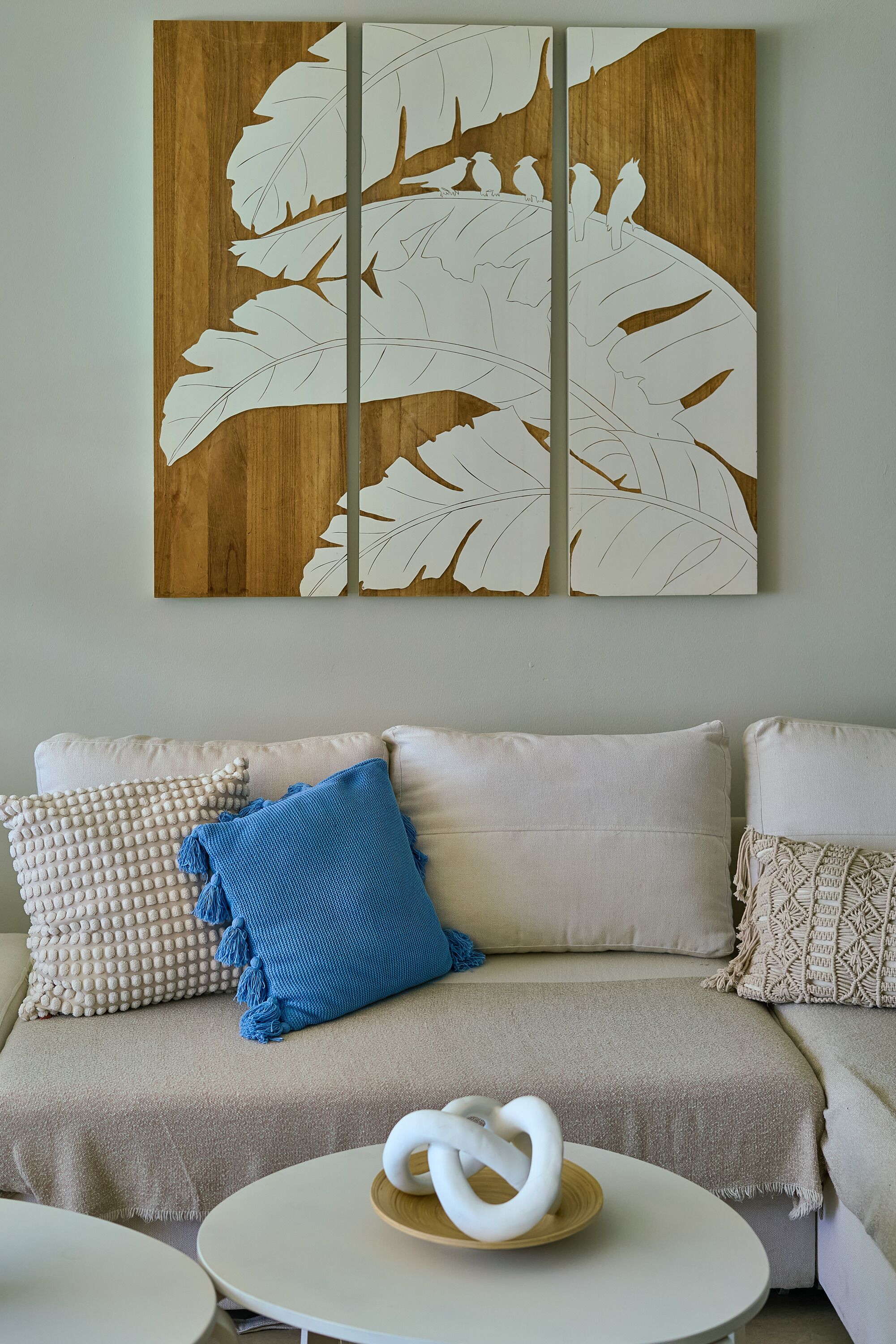 Una sala de estar con un sofá color beige, un cojín azul y una obra de arte abstracta blanca en la pared.