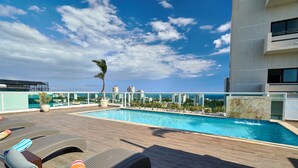 A rooftop pool with lounge chairs, a palm tree, and a city skyline in the background. - The LUX Natural Art Downtownsd Pool Gym 2BR (Santo Domingo)