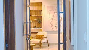 A hallway with a wooden door, a white chair, a small table, and a wall with abstract art. - The LUX Natural Art Downtownsd Pool Gym 2BR (Santo Domingo)