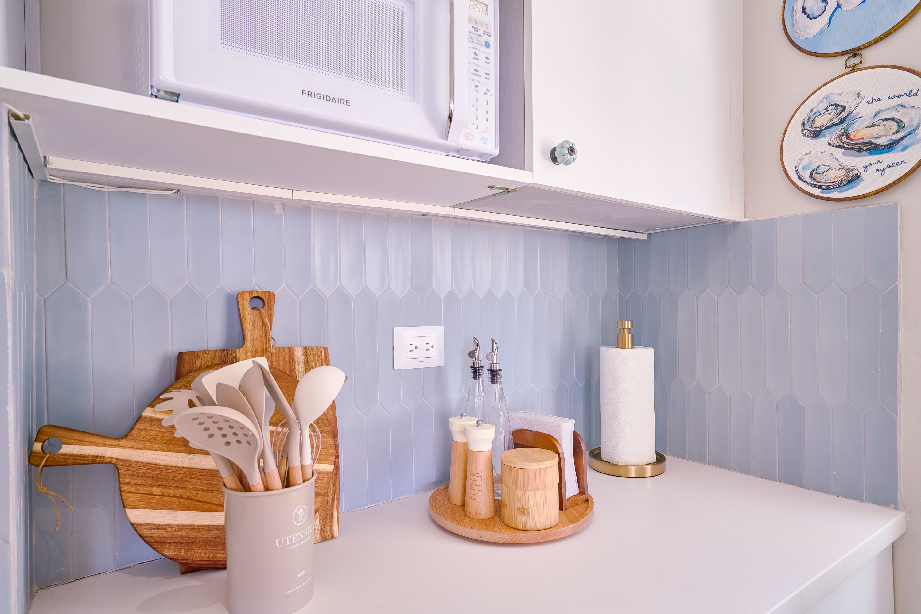 Een keuken met een magnetron, houten snijplank, keukengerei en een dienblad met flessen en een kaars.