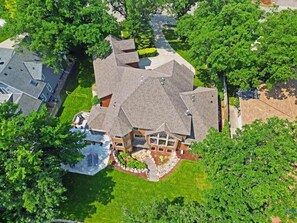 A large, multi-story house with a complex roof structure, surrounded by lush greenery and a well-maintained lawn.