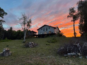 Une maison avec un grand jardin à l’avant, une terrasse en bois et une vue dégagée sur le ciel.