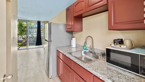 A kitchen with a stainless steel refrigerator, granite countertop, red cabinets, a microwave, and a sink. - Private Balcony • Walk to Beach + Downtown (Hilo)