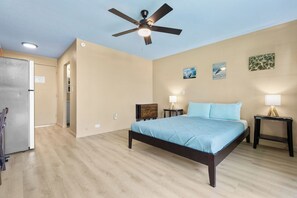 A bedroom with a bed, a ceiling fan, a refrigerator, and a nightstand with a lamp.
