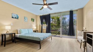 A bedroom with a bed, bedside tables, a ceiling fan, a desk, and a sliding glass door leading to a balcony. - Private Balcony • Walk to Beach + Downtown (Hilo)