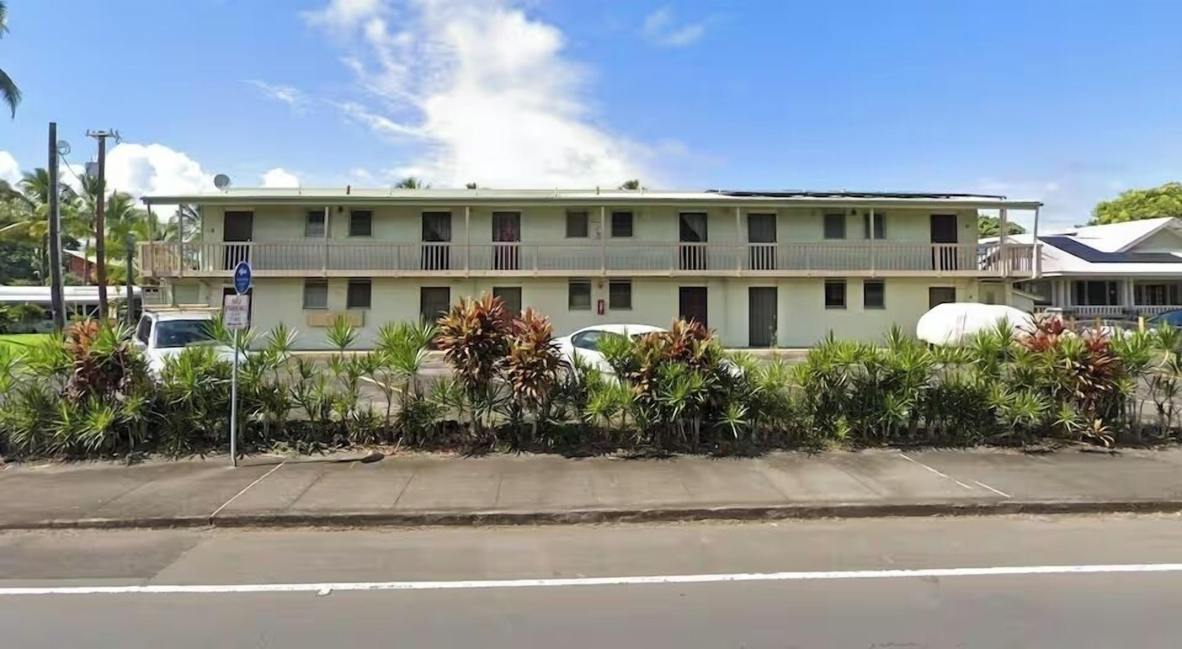 A two-story motel with a flat roof, multiple balconies, and a parking area with cars.