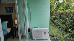 A room with a bed, a nightstand, a lamp, and a sliding glass door leading to a balcony with a small air conditioning unit.