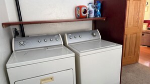 Un área de lavandería con dos lavadoras y secadoras de carga frontal blancas, un estante con productos de limpieza y una puerta de madera.
