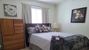 Un dormitorio con una cama, un aparador de madera, una mesita de noche, una lámpara y dos cuadros de flores colgados en la pared.