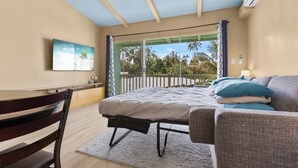 Ein Schlafzimmer mit Bett, Sessel, Schreibtisch, Deckenventilator und Blick auf Palmen durch eine Schiebetür.