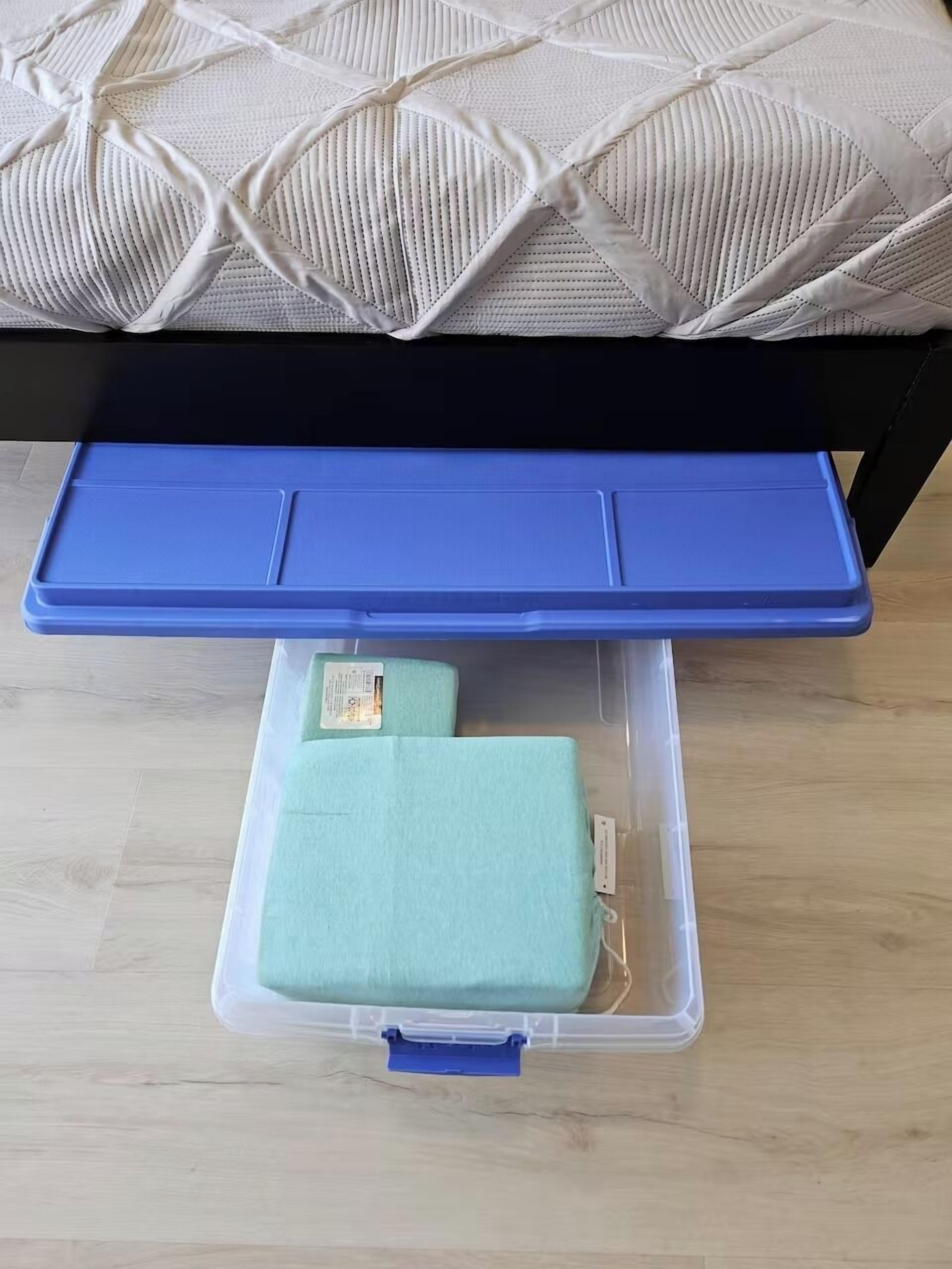 A bed with a patterned white cover, a blue bedside table, and a storage box with a green item inside.