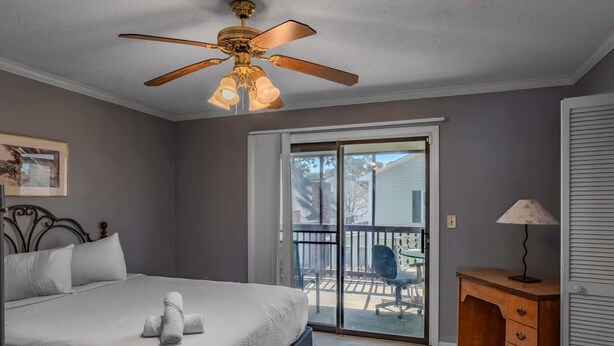 Une chambre à coucher comprenant un lit, un ventilateur de plafond, un bureau avec des tiroirs, une chaise et une porte-fenêtre coulissante donnant sur un espace extérieur.