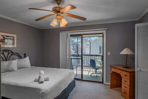 Une chambre à coucher comprenant un lit, un ventilateur de plafond, un bureau avec des tiroirs, une chaise et une porte-fenêtre coulissante donnant sur un espace extérieur.