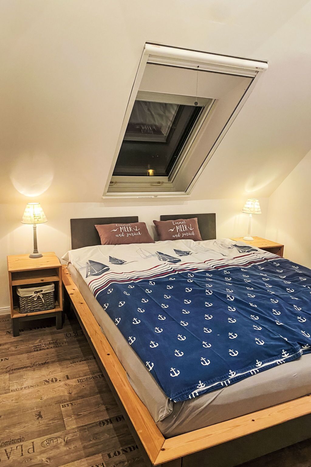 Un dormitorio con tragaluz, una cama con manta azul, dos lámparas de noche y una mesita de noche con lámpara.