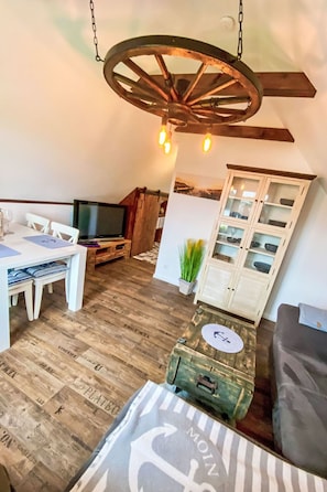 A loft-style room with a wooden floor, a dining table with chairs, a television, a cabinet with glass doors, and a hanging light fixture. - Apartment AHOI by Interhome (Norddeich (Dithmarschen))