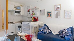 Una cucina moderna con mobili bianchi, un forno a microonde e una macchina da caffè. C'è un tavolo da pranzo con bicchieri e una ciotola rossa. Un divano blu con cuscini decorativi è appoggiato al muro.