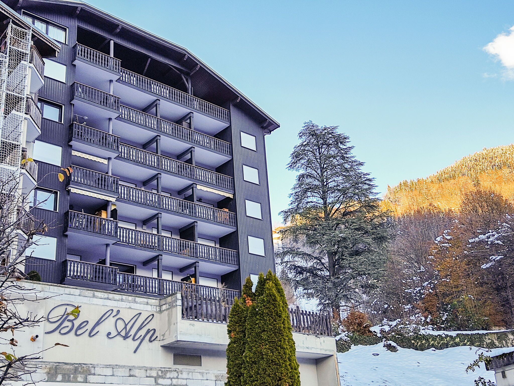 Un hotel de varios pisos con balcones, un letrero que dice 'Bel Alp' y un paisaje nevado al fondo.