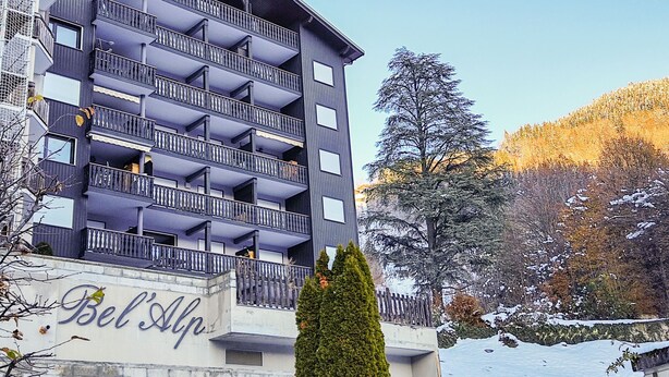 Un hotel a più piani con balconi, un cartello che riporta 'Bel Alp' e un paesaggio innevato sullo sfondo.