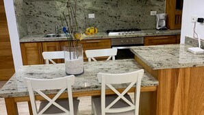 Une cuisine dotée de plans de travail en granit, de chaises blanches, d’un îlot central, de meubles en bois et d’une hotte de cuisine en acier inoxydable.