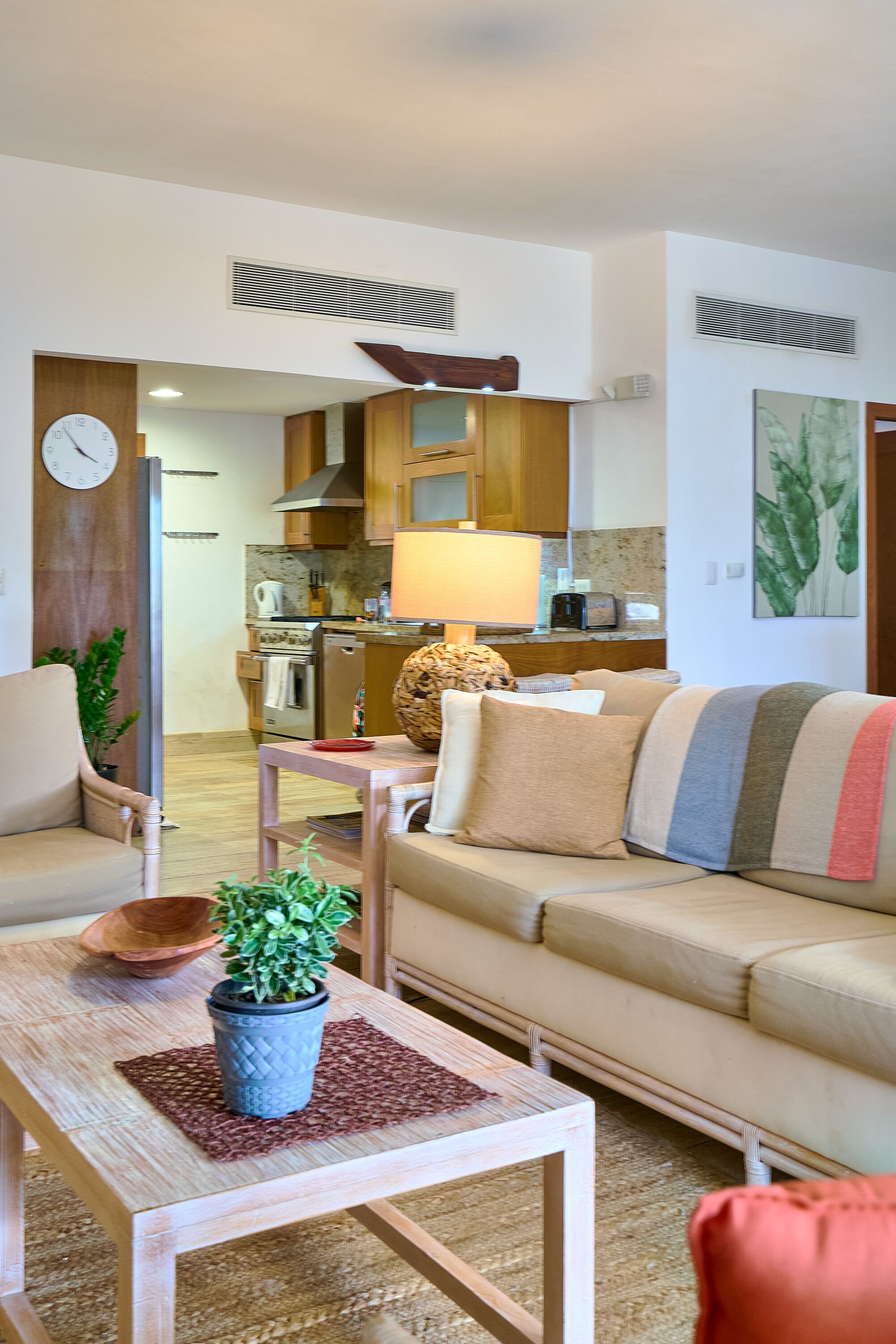 Un salon avec un canapé, une table basse ornée d’une plante, une table d’appoint en bois, une cuisine en arrière-plan et une horloge au mur.
