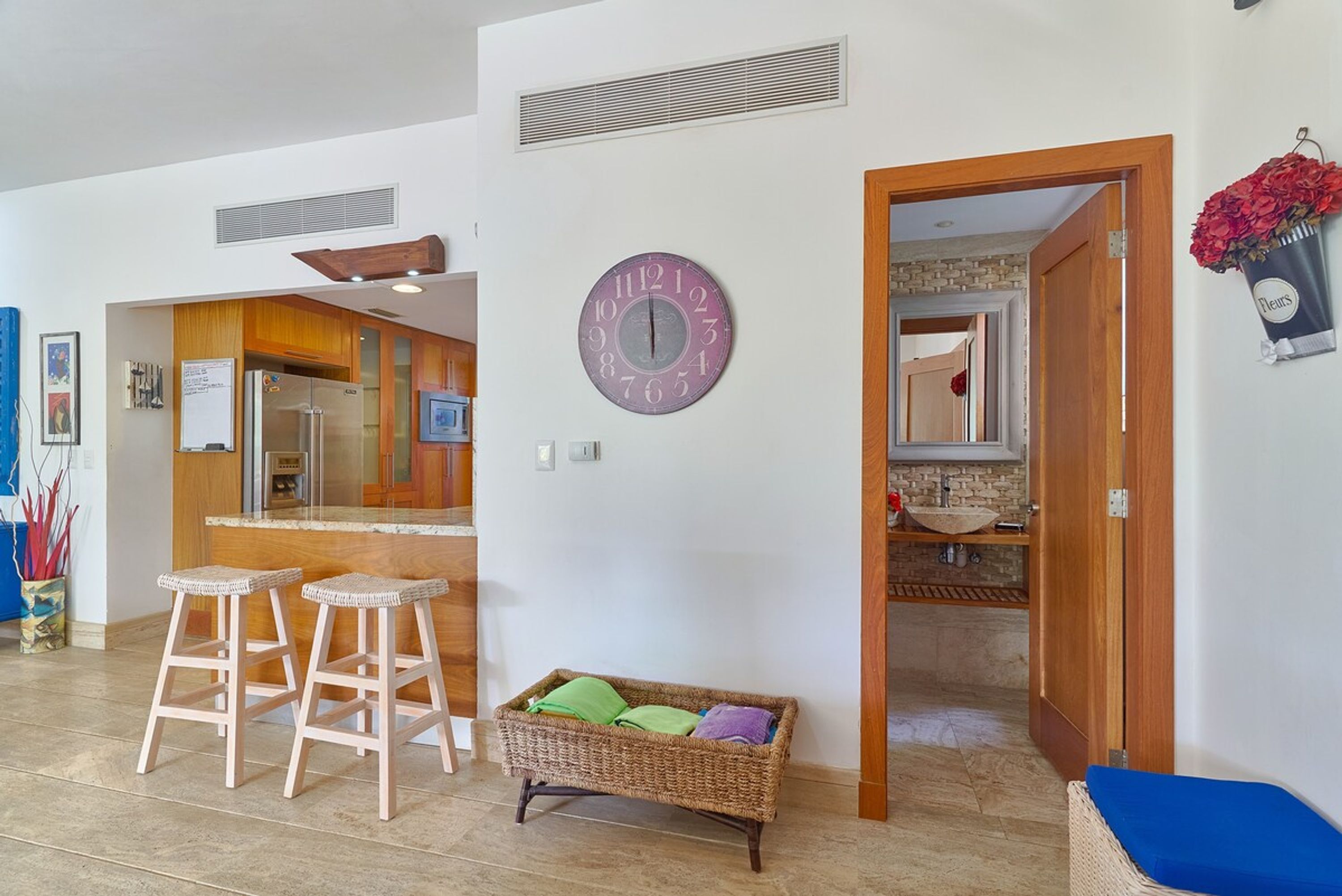 Une cuisine moderne avec un îlot central, des tabourets en bois, un panier en osier et une horloge murale.