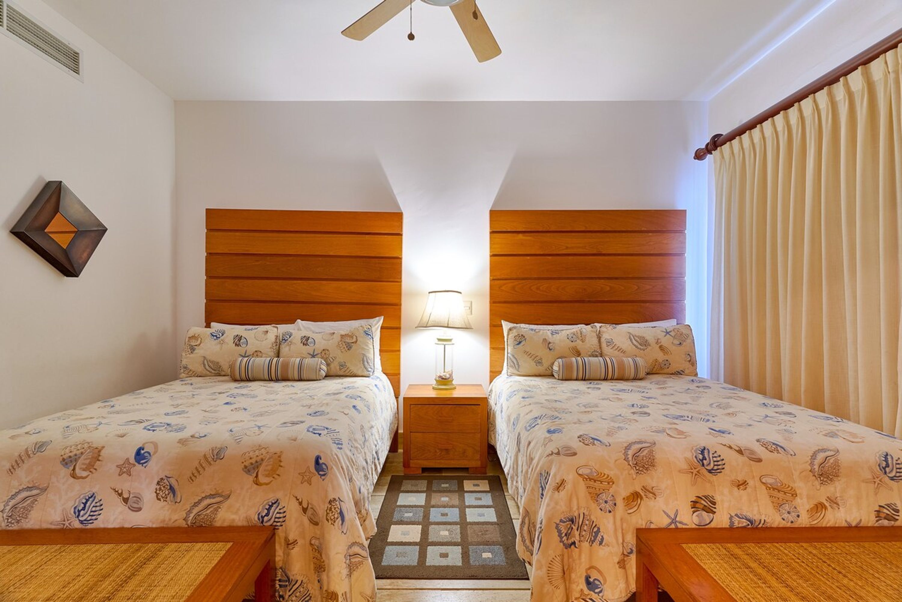 A room with two beds, a wooden headboard, a nightstand with a lamp, a patterned rug, and a ceiling fan.