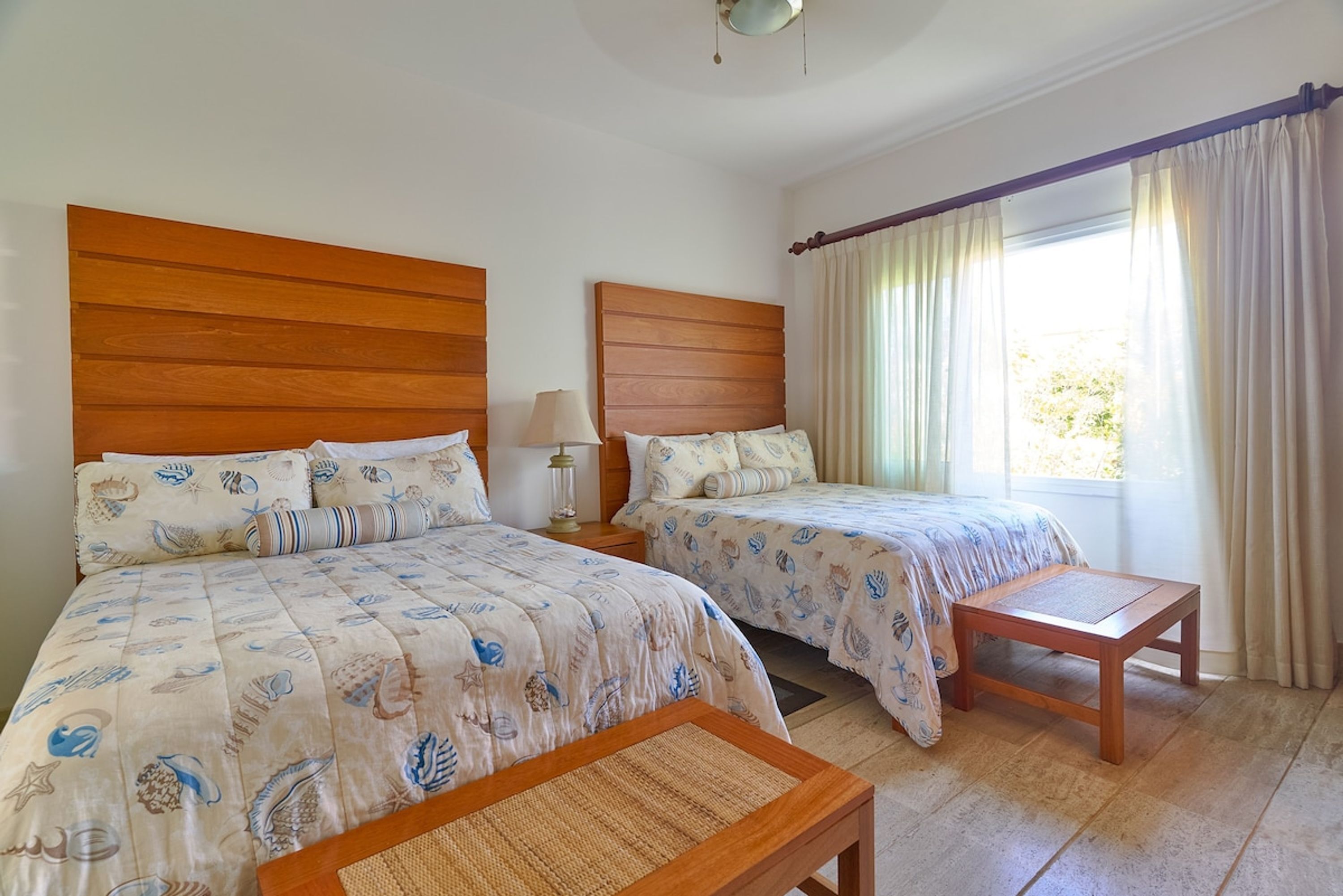 A room with two beds, a wooden headboard, a bedside table with a lamp, and a window with curtains.