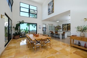 A spacious dining area with a large wooden table and chairs, a kitchen with white cabinets, and large windows allowing natural light. - Beach Mountain Villa Terrenas Pool BBQ 6BR (Las Terrenas)