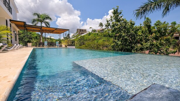 A clear swimming pool with a wooden deck and lounge chairs, surrounded by lush greenery and a clear sky. - Beach Mountain Villa Terrenas Pool BBQ 6BR (Las Terrenas)