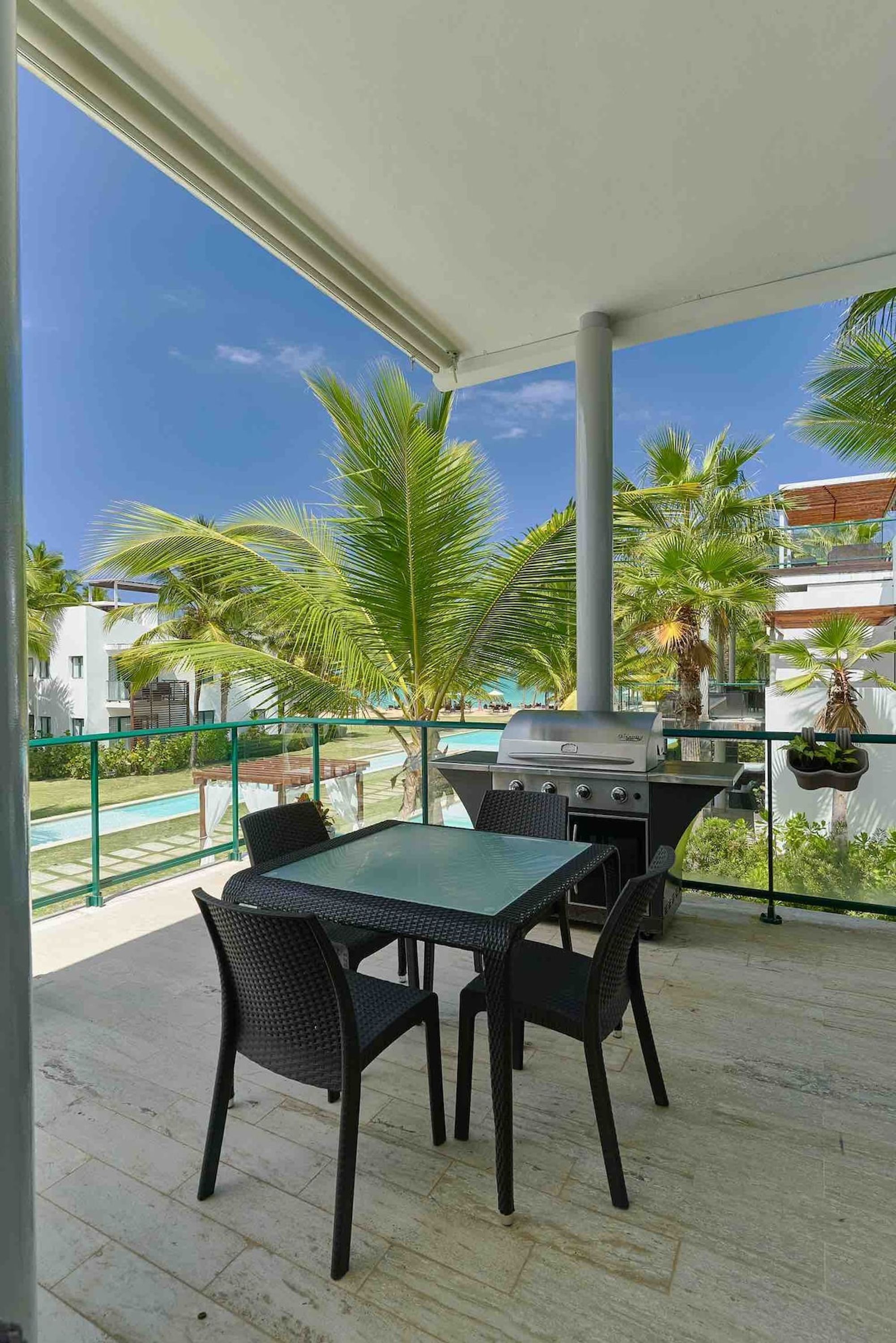 Une terrasse extérieure couverte, équipée d’un barbecue, d’une table à manger et de chaises, avec vue sur une piscine et des palmiers.