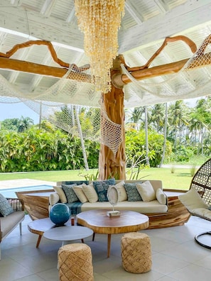 A tropical outdoor lounge area with a wooden column, woven ceiling, and a view of lush greenery. - The Palms View Playabonita Beachfront Pool BBQ (Las Terrenas)