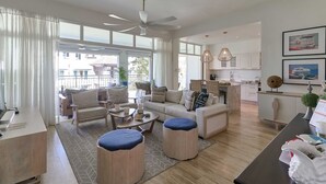 Amplia sala de estar con sofá, otomanes, una mesa de centro y una mesa de comedor. Se aprecia una cocina al fondo con gabinetes blancos y una lámpara colgante.