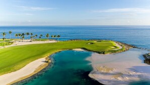 Un campo de golf costero con vistas despejadas al océano, zonas arenosas y palmeras.