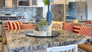 Una cocina moderna con encimeras de granito, una isla central, cojines estampados en rojo y blanco e iluminación colgante.