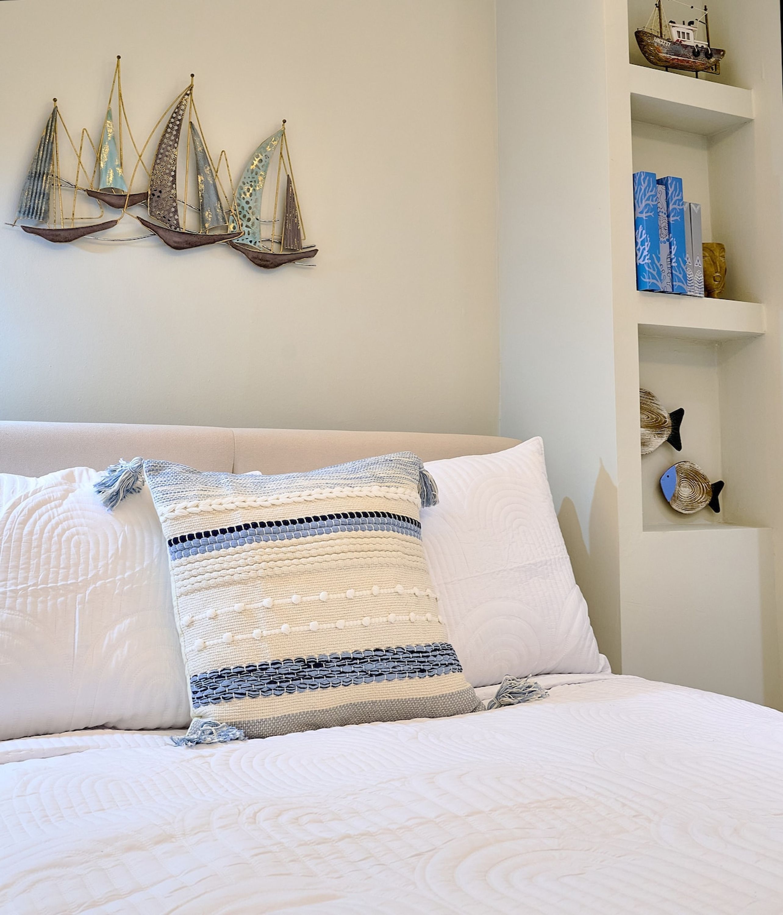 A bed with white bedding, a decorative pillow, and a wall with a shelf holding a ship model and a fish decoration.