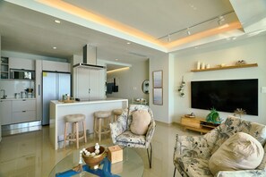 A modern living room with a sofa, a chair, a TV, and a kitchen area in the background. - Marbella LUX Essence Beachfront Pool 11thfloor (Guayacanes)
