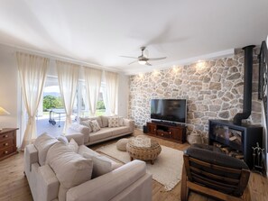 A living room with a stone wall, a flat-screen TV, a sofa, a wooden bench, and a fireplace. - Vacation home Olivia by Interhome (Šestanovac)