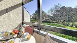 A balcony with a table set for breakfast, including fruits and drinks, overlooking a garden with trees and a lawn. - Les Jardins de la Plage by Interhome (Carnac)