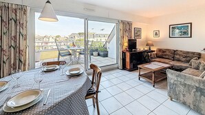 A living room with a dining area, a sofa, a television, and a balcony with a view of buildings. - Le Hoche by Interhome (Quiberon)
