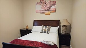 A neatly made bed with a wooden headboard, a nightstand with a lamp, and a wall-mounted picture of cherry blossoms. - Enjoy a piece of home in a cozy inviting space. (Waynesboro)