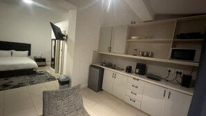 Un dormitorio con cama, mesita de noche y una silla. La habitación tiene una kitchenette con microondas, cafetera y armarios.