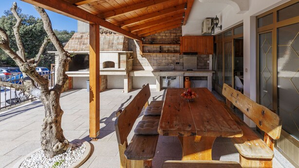 A covered outdoor dining area with a wooden table and benches, a brick fireplace, and a kitchen area with cabinets and a refrigerator. - Milanković by Interhome (Vrsar)