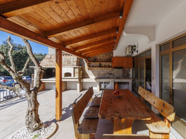 A covered outdoor dining area with a wooden table and benches, a brick fireplace, and a kitchen area with cabinets and a refrigerator. - Milanković by Interhome (Vrsar)