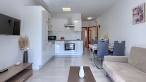 A modern living room with a sofa, a dining table with chairs, a kitchen area with cabinets and appliances, and a TV mounted on the wall. - Milanković by Interhome (Vrsar)